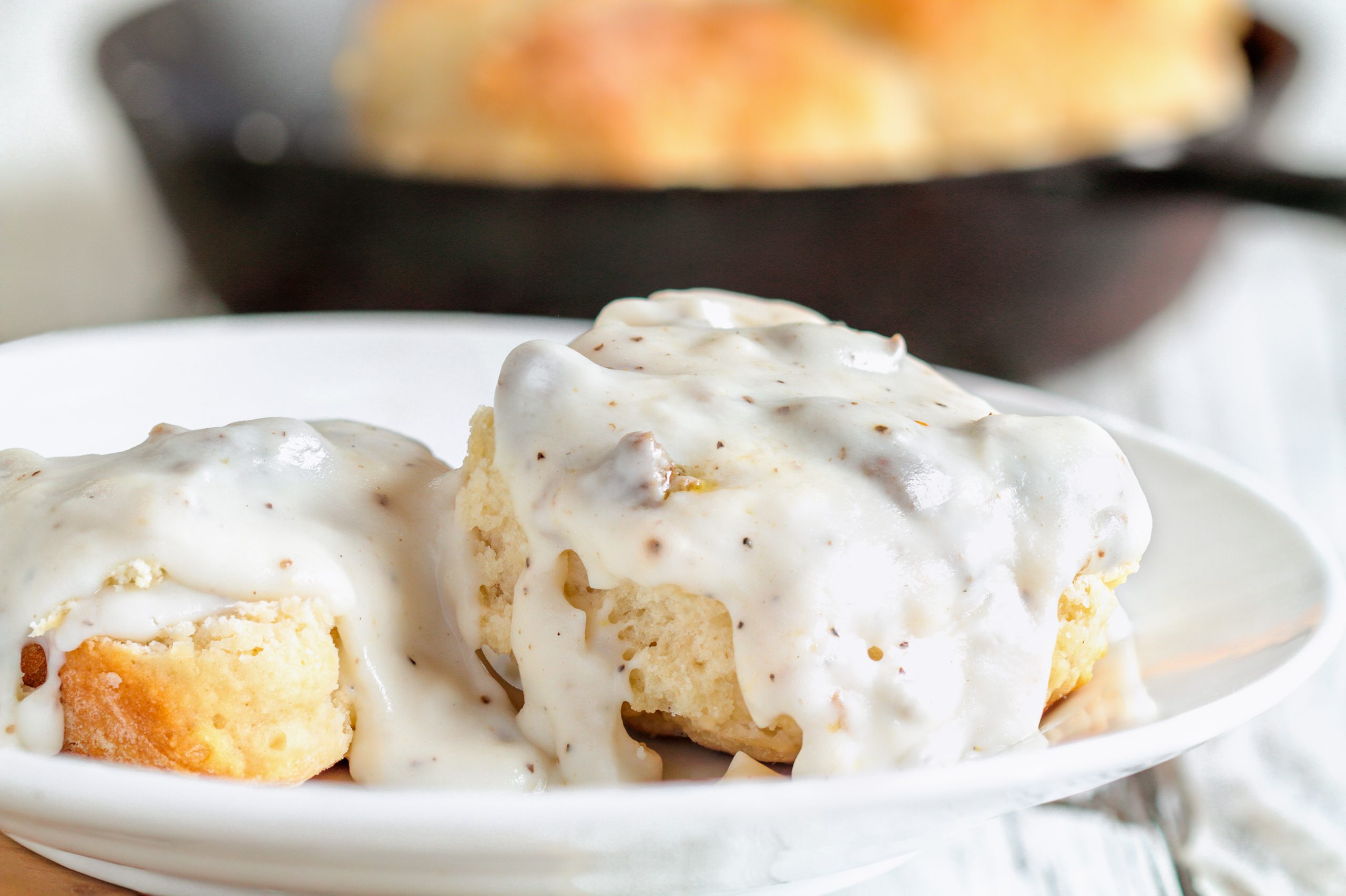 american biscuits from scratch covered with thick white sausage gravy. selective focus with cast iron skillet / pan in the background over a white table.
